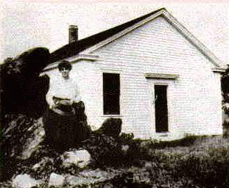 Mrs. Marie B. McNitt seated on rock before Palmer schoolhouse voted in 1762